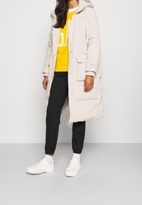 Giaccone imbottito beige chiaro con cappuccio, chiusura con zip, tasche laterali e orlo con coulisse. Indossato sopra una felpa gialla e pantaloni scuri con motivi.