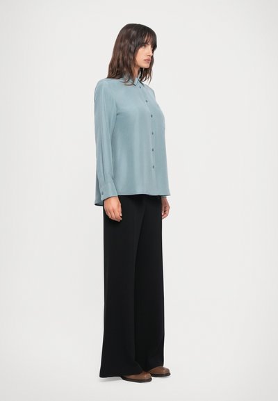 Light blue long-sleeve shirt with a button front, pocket, and relaxed fit paired with black wide-leg trousers and brown flats.