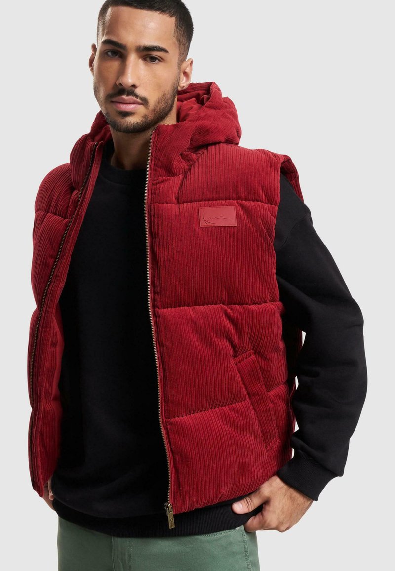 Rød kordfløyels puffer vest med hette, med glidelås og sidelommer; strukturert stoff og et rektangulært logo-patch foran.