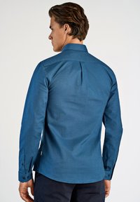 Langarm overhemd in donkerblauwe stof met een gladde textuur, met een rugplooitje en knoopmanchetten.