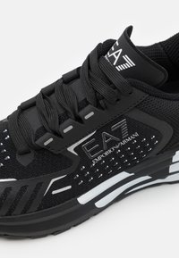 Musta urheilukenkä verkkopäällisellä, vahvistetulla varpaalla ja nauhoitusjärjestelmällä. Kenkässä on valkoiset yksityiskohdat ja brändäys sekä teksturoitu, pehmustettu pohja.