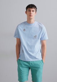 Ljusblå t-shirt i bomull med korta ärmar, prydd med broderade sol-, måne- och fågeldesigner. Dagens outfit är kombinerad med turkosa shorts.