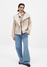 Beige wolmix dubbele knoopsluiting jas met brede revers, gecombineerd met een lichte trui en blauwe high-waisted uitlopende jeans. Bruine schoenen maken de look compleet.