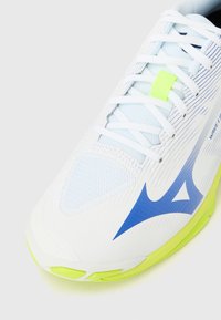 Λευκό αθλητικό παπούτσι με αεριζόμενο επάνω μέρος από δίχτυ, μπλε λεπτομέρειες, πράσινο ταμπελάκι κορδονιού και υφή στη σόλα για σταθερότητα.