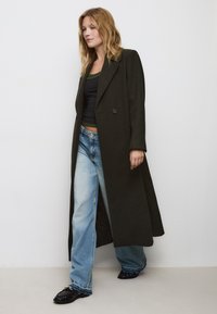 Cappotto lungo di colore verde scuro con colletto a punta, chiusura a singolo bottone, abbinato a una canotta nera e a jeans a gamba larga di colore blu chiaro.
