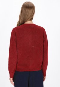 Vrouw met lang lichtbruin haar in een donkerrode gebreide trui met subtiele glitters en donkere broek, gezien vanuit de achterkant.