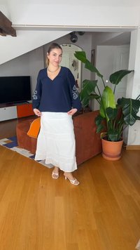 Γυναίκα με σκούρα μαλλιά δεμένα πίσω φοράει μπλε μπλούζα και λευκή φούστα, στέκεται σε ξύλινο πάτωμα κοντά σε πορτοκαλί καναπέ και μεγάλο φυτεμένο φυτό.