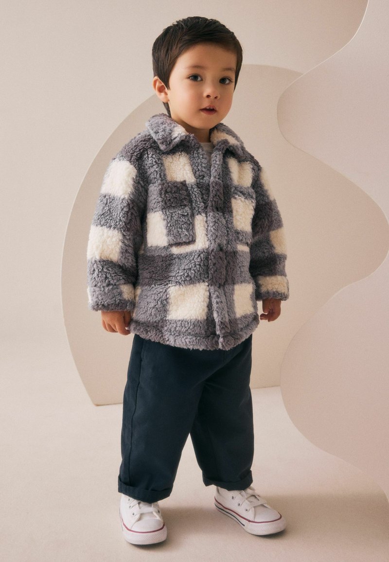 Jeune enfant portant une veste en polaire à carreaux gris et blancs, un pantalon sombre et des baskets blanches, se tenant à l'intérieur devant un fond beige.