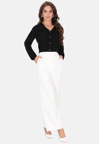 Cardigan negro de punto con cuello en V y cierre de botones, combinado con pantalones blancos de pierna ancha. Modelo posando con las manos en los bolsillos.