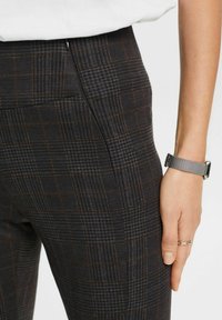 Gros plan d'une personne portant un pantalon à carreaux gris foncé et un haut blanc, avec une montre à maille argentée et une bague à maillons sur la main droite.