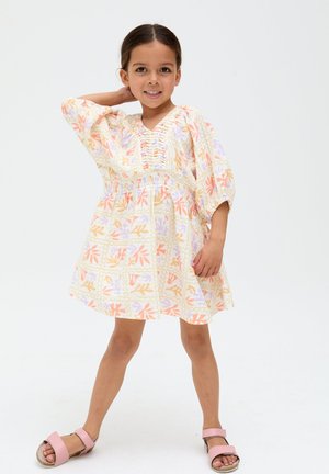 Lächendes Mädchen steht mit einer Hand hinter dem Kopf, trägt ein pastellfarbenes Blumenkleid und rosa Sandalen vor weißem Hintergrund.