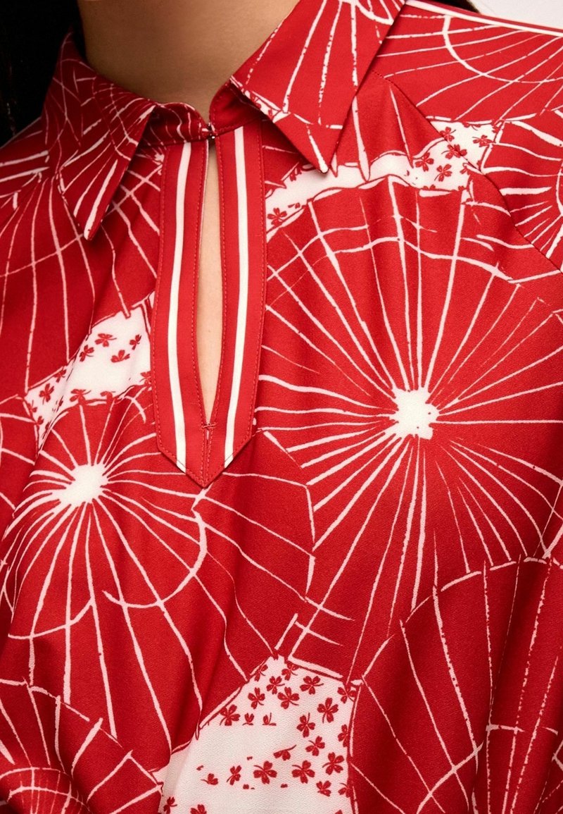 Chemise rouge avec motifs radiaux blancs de parapluie et détails floraux, comportant une petite fente verticale à l'encolure et un col pointu.
