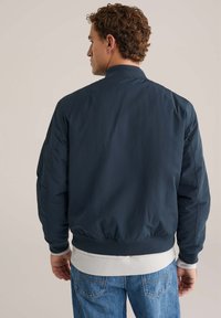 Giubbotto bomber navy con una texture liscia, polsini a coste e banda inferiore bianca. Progettato con colletto alto e tasche laterali.