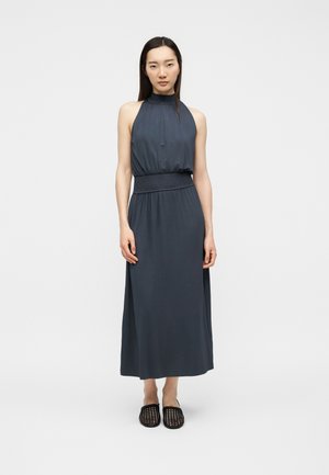 Kvinde iført en ærmeløs, mørkegrå kjole med høj hals og indsnævret talje samt sorte vævede slip-on sko, stående mod en lys baggrund.
