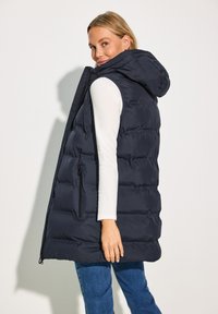 Gilet en duvet bleu marine avec capuche, présentant un design matelassé, deux poches latérales zippées et une silhouette fluide. La texture semble lisse.