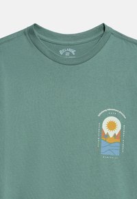 Πράσινο μπλουζάκι με στρογγυλή λαιμόκοψη και μικρό γραφικό με ήλιο, βουνά, φοίνικα και νερό, με ετικέτα "Billabong Adventure Division" στο στήθος.