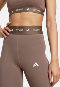 Braune sportliche Leggings mit einem breiten Bund, der die "TECHFIT"-Marke in Weiß trägt. Glatte Textur mit schmalem Schnitt und Seitennaht.
