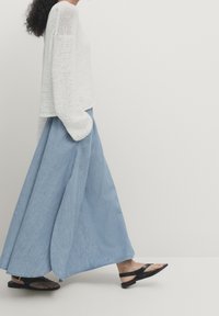 Donna che cammina, indossando pantaloni a gamba larga blu chiaro, un maglione bianco lavorato a maglia largo e sandali flat neri, contro uno sfondo bianco semplice.