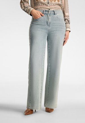 Woman wearing light blue wide-leg jeans with a patterned long-sleeve blouse and brown pointed-toe heels, standing with one hand in pocket.