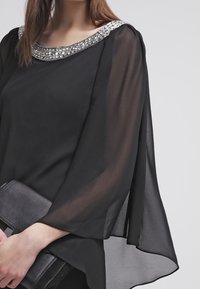Blouse noire transparente avec un col orné de bijoux. Caractérisée par des manches fluides et un tissu délicat. Associée à une pochette noire.