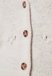 Cardigan lavorato a maglia color crema con tessuto testurizzato, cinque bottoni marroni e leggeri segni di usura vicino alla placca dei bottoni.