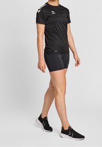 Musta urheilupaita lyhyillä hihoilla, jossa on logo ja valkoiset chevron-tyyliset yksityiskohdat hartioissa, yhdistettynä mustiin shortseihin ja urheilukenkiin.