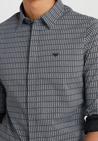 Svart och vit randig skjorta med knapp-down krage och liten logotyp. Tyget visar ett repetitivt textmönster. Texturerad, figurnära design.