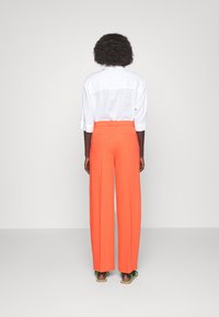 Pantaloni arancioni a gamba larga con vita alta, dotati di passanti per cintura e due tasche posteriori, abbinati a una camicia bianca dal taglio morbido.