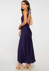 Mujer vistiendo un vestido sin mangas, de color azul marino y espalda descubierta, con pendientes dorados, sosteniendo un clutch beige y usando tacones de plataforma beige, de pie contra un fondo liso.