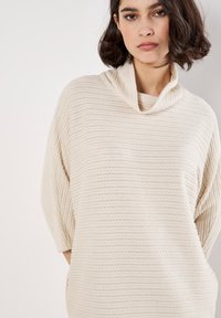 Maglione color crema con collo a scaldacollo, dotato di un motivo testurizzato a righe e maniche a tre quarti, realizzato in un materiale morbido e flessibile.