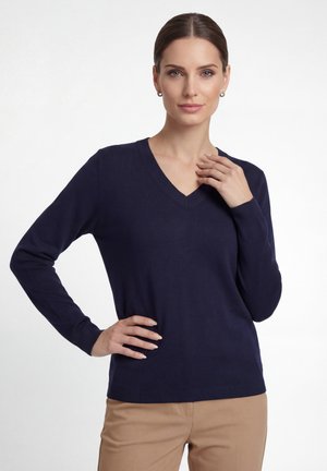 Kvinde med brunt hår iført sølvfarvede øreringe, en marineblå V-hals trøje og beige bukser, poserer med hånden på hoften og nær kravebenet.