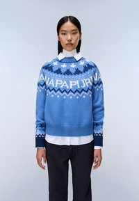 Maglione blu lavorato a maglia con motivi geometrici in blu scuro e bianco, collo rotondo, tessuto testurizzato, maniche lunghe, indossato sopra una camicia bianca.