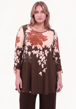 Kvinde iført brun tunika med fersken- og rustfarvede blomsterprint og trekvartærmer bundet ved manchetterne, kombineret med mørke bukser.