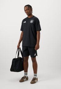 Μαύρη αθλητική μπλούζα με λογότυπο, αντίστοιχες μαύρες σορτς, και καφέ παπούτσια. Τα αξεσουάρ περιλαμβάνουν μια μαύρη τσάντα αθλητισμού. Οι ριγωτές κάλτσες ολοκληρώνουν την εμφάνιση.