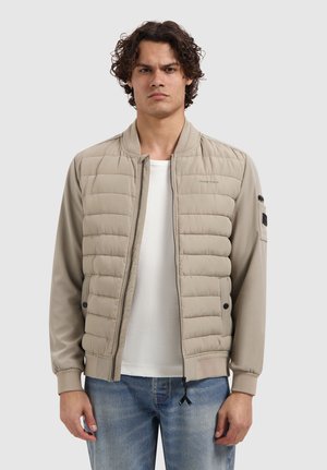 Jonge man met krullend haar, gekleed in een beige doorgestikte bomberjack over een wit shirt en blauwe spijkerbroek, staand tegen een effen achtergrond.