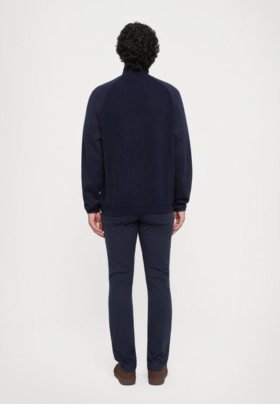 Hanorac bleumarin închis cu guler înalt, având mâneci raglan și material texturat. Asortat cu pantaloni corespunzători și încălțăminte maro.