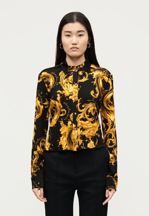 Kobieta ubrana w czarną koszulę zapinaną na guziki z wyrazistymi żółtymi barokowymi zawijasami oraz czarne spodnie, stojąca na jednolitym jasnym tle.