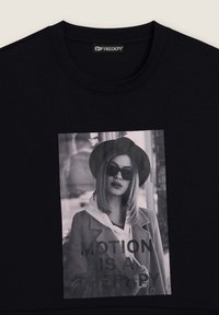 Maglietta nera con una foto in scala di grigi di una donna che indossa occhiali da sole, cappello e cappotto, con la scritta "IL MOVIMENTO È UNA TERAPIA" nella parte anteriore della maglietta.