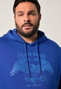 Sudadera azul hecha de tela suave, con un gráfico de alas de águila y detalles en texto. Los cordones en la capucha realzan el diseño.