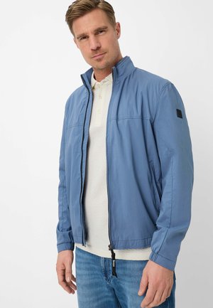 Man draagt een lichtblauw jack met rits over een wit poloshirt en blauwe spijkerbroek, staand tegen een effen witte achtergrond.