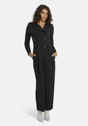 Zwarte jumpsuit met lange mouwen, reverskraag, dubbele knoopsluiting, zijzakken en een strikriem in de taille. De silhouet is getailleerd en slank.
