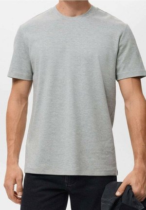 Mann trägt ein schlichtes hellgraues Kurzarm-T-Shirt und dunkle Hosen, hält eine dunkle Jacke in seiner rechten Hand.