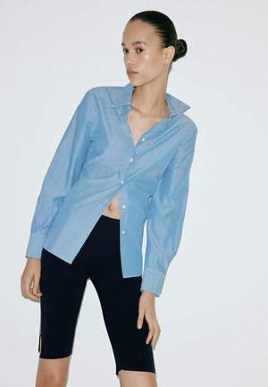 Femme aux cheveux plaqués en arrière porte une chemise rayée bleue partiellement ouverte et un short de cyclisme noir, posant devant un fond uni.