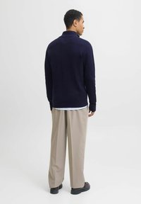 Jūrinės spalvos megztinis su aukštu apykakliu, vilkimas ant balto marškinio, derinamas su šviesiais plačiais kelnėmis ir tamsiais batais. Vaizdas iš galo.