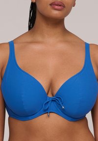 Blauw gewatteerd bikini-topje met een diepe halslijn, verstelbare bandjes en een knoopdetail aan de voorkant. Gemaakt van zachte, rekbare stof.