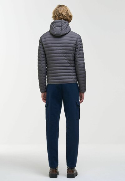 Personne aux cheveux châtain clair portant une veste matelassée grise à capuche, un pantalon cargo bleu marine et des chaussures marron, debout de dos sur un fond uni.