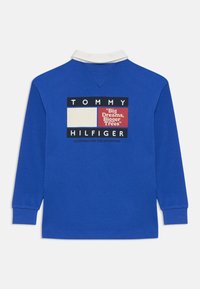 Polo bleu à manches longues avec un col blanc. Doté d'un grand logo imprimé au dos avec un bloc rouge et du texte.