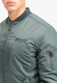 Green bomber jacket made of lightweight fabric, featuring a ribbed collar, zip closure, side pockets, and a logo embroidered on the chest.