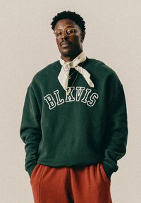 Mann mit Brille, der einen dunkelgrünen BLKVIS-Pullover, einen weißen gemusterten Schal, goldene Halsketten und rostrote Hosen trägt, steht mit den Händen in den Taschen.