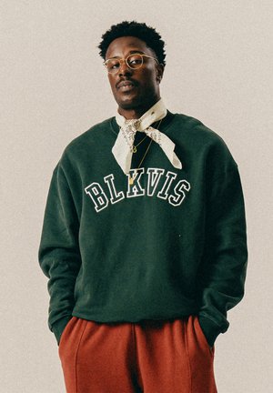 Mann mit Brille, der einen dunkelgrünen BLKVIS-Pullover, einen weißen gemusterten Schal, goldene Halsketten und rostrote Hosen trägt, steht mit den Händen in den Taschen.